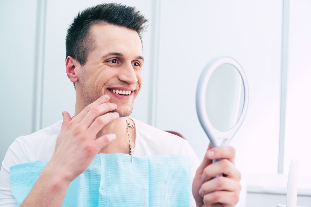 Man in dental office examining his smile in handheld mirror, wearing light blue shirt.