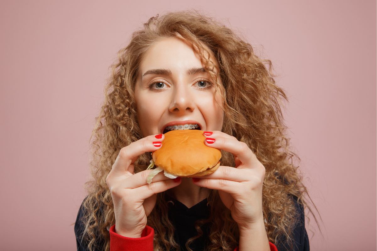 Woman with curly hair biting into a burger against pink background, displaying healthy teeth.