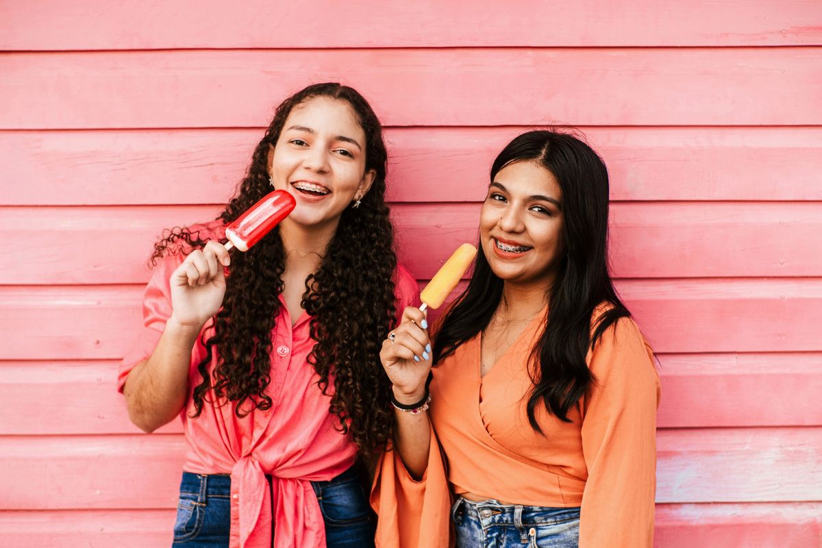 Two young women smiling and holding lollipops against a pink wooden wall background.