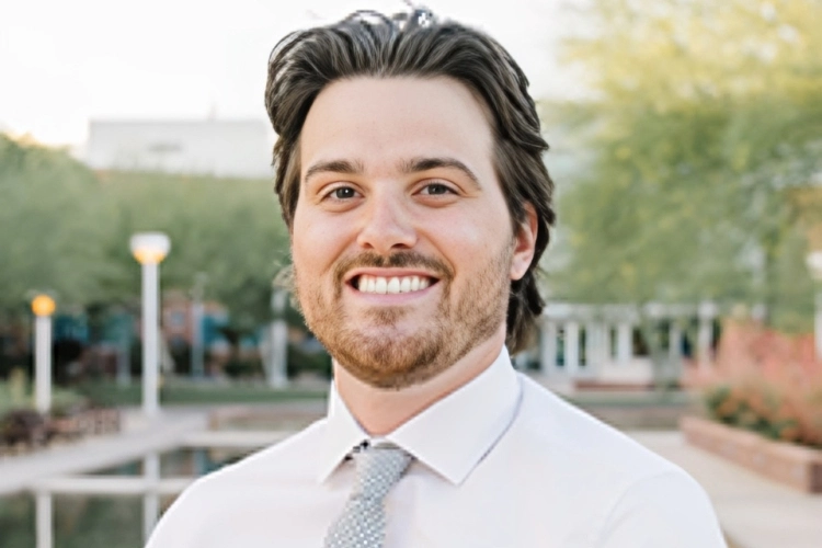 Professional man in light blue shirt and tie smiling outdoors near water fountain.