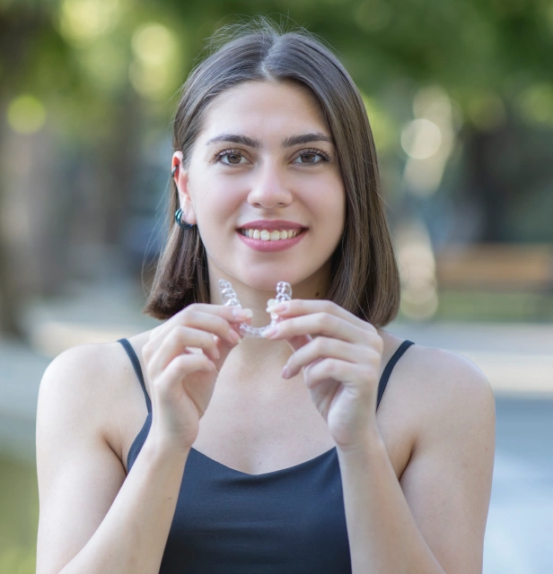 Young woman holding orthodontic aligners outdoors, smiling at camera in natural daylight setting.
