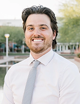 Professional headshot of smiling man in white shirt and tie outdoors.