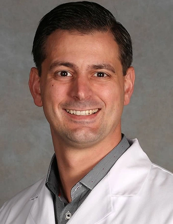Professional headshot of male dentist wearing white coat against gray background.