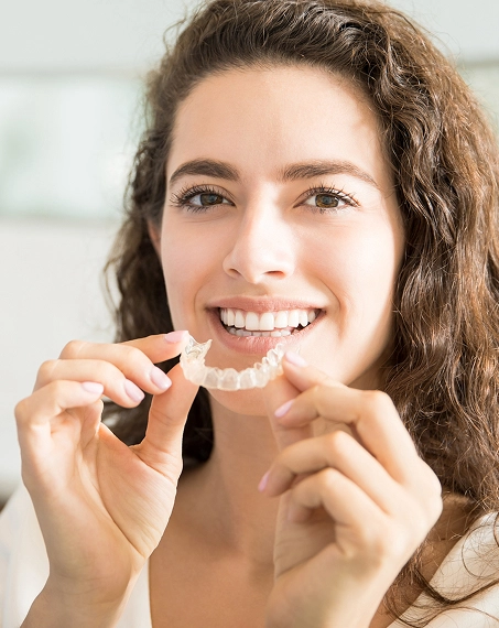 Woman smiling while holding clear orthodontic aligners in bright dental office setting.