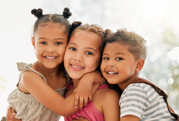 Three smiling children hugging together in a bright, indoor setting.