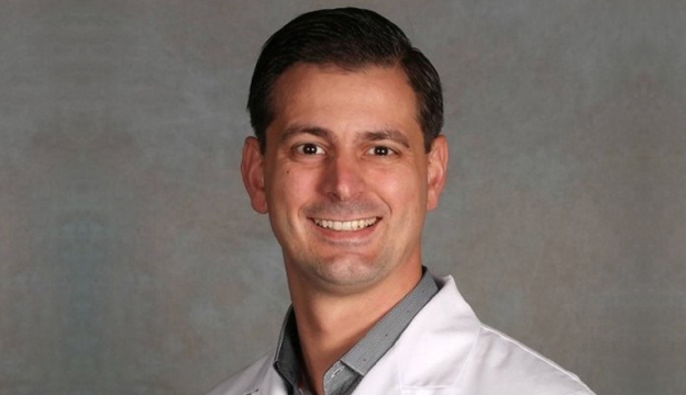 Professional headshot of male dentist in white coat smiling against gray background.