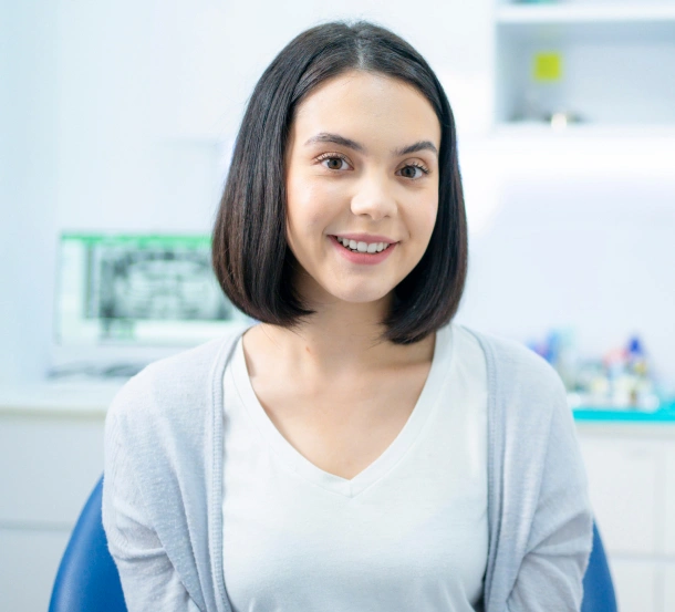 Young woman smiling in dental office with treatment chair and supplies visible in background.