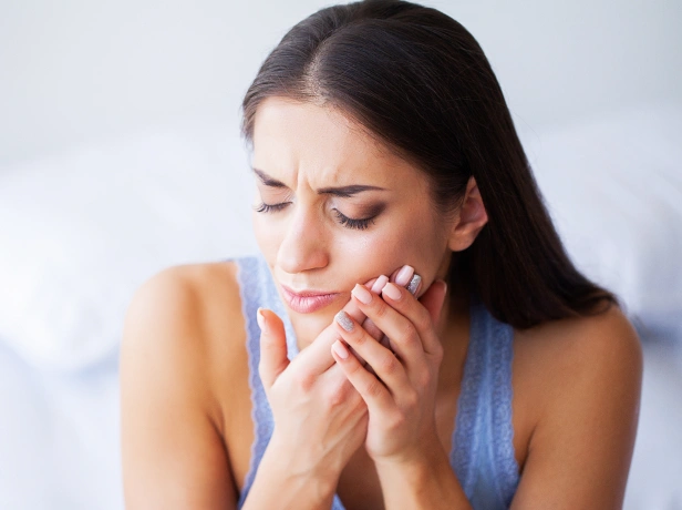 Woman experiencing tooth pain, holding cheek with hands in distress against white background.