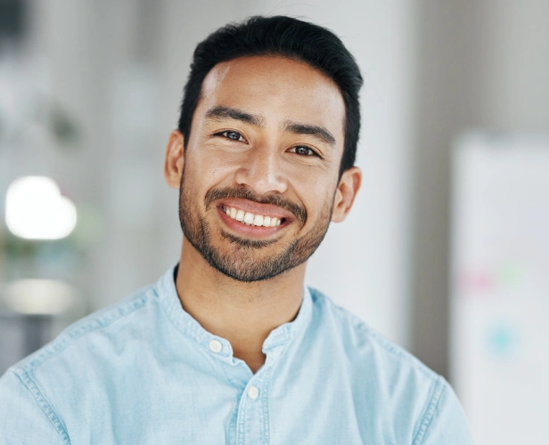 Smiling man in light blue shirt against blurred office background, displaying healthy teeth and friendly expression.