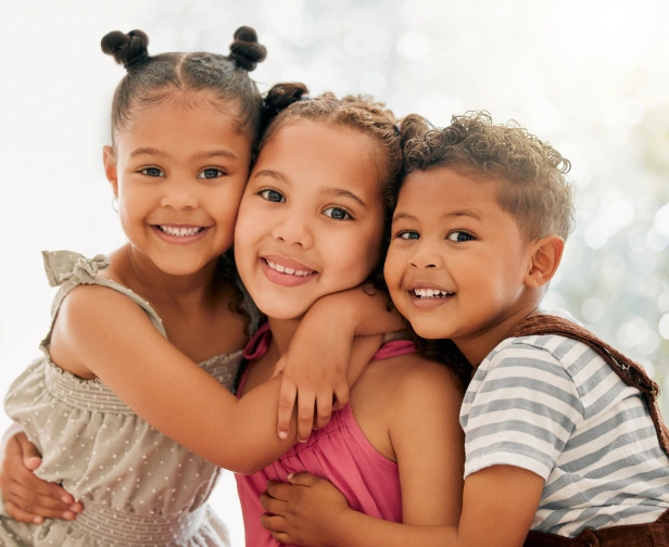 Three smiling children hugging together in a bright, cheerful studio setting.
