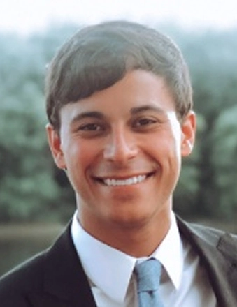 Professional headshot of smiling man in business suit with outdoor blurred background.