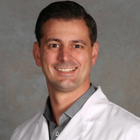Male dental professional in white coat smiling against gray background, headshot portrait.