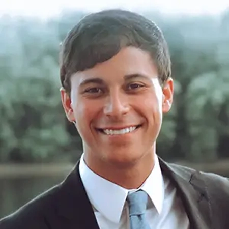 Professional headshot of smiling man in business suit against blurred outdoor background.