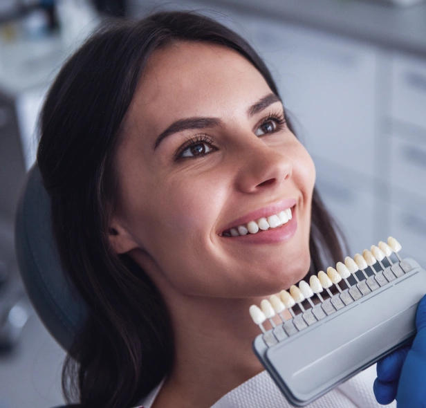 Smiling woman holding teeth shade guide in dental office setting.