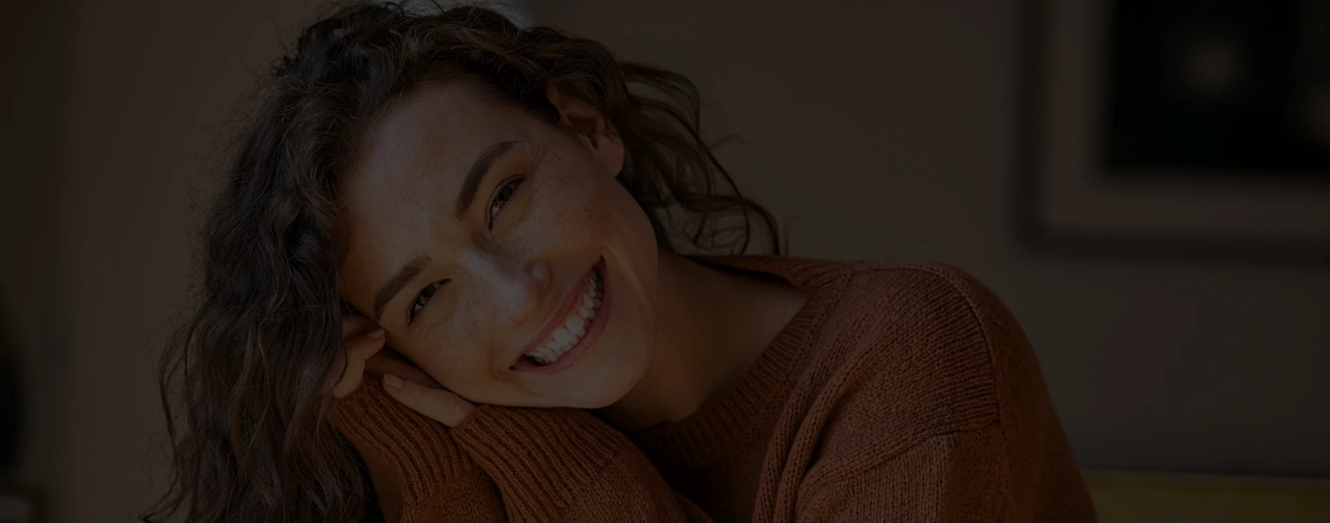 Woman with dark curly hair smiling warmly in indoor setting, wearing brown shirt.