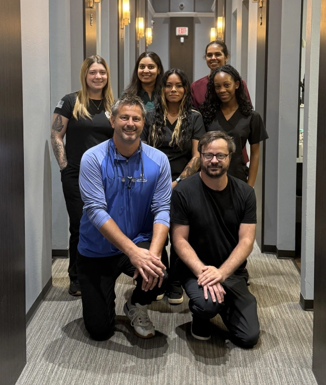 Dental team of eight professionals posing together in a modern office hallway.