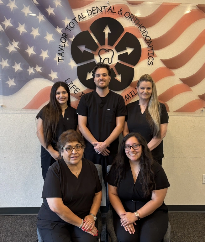 Five dental team members in black scrubs posing together in front of American flag and dental office logo.